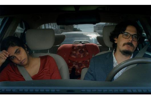 still from film; a man and woman driving in car, looking unhappy